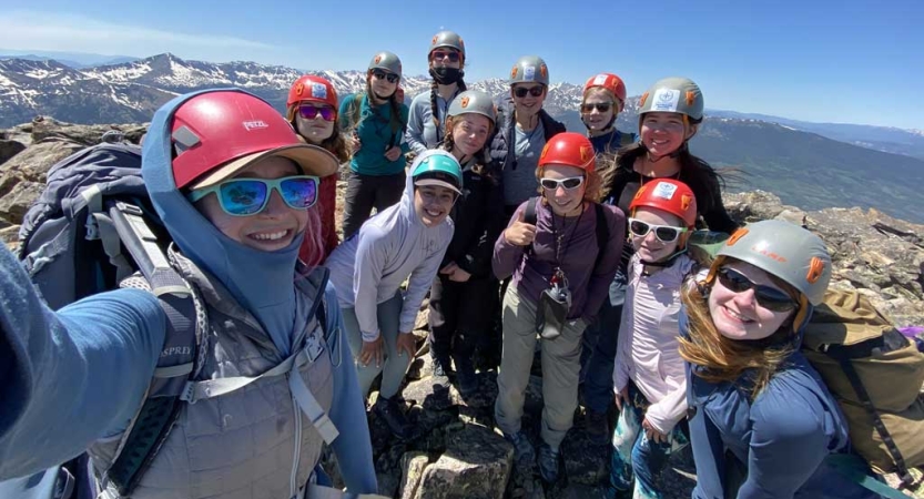 a group of people wearing helmets stand on the summit of a mountain, high above the mountainous landscape below. 